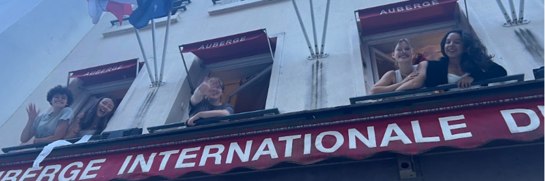 A few students lean out a window of a hotel and wave to those below