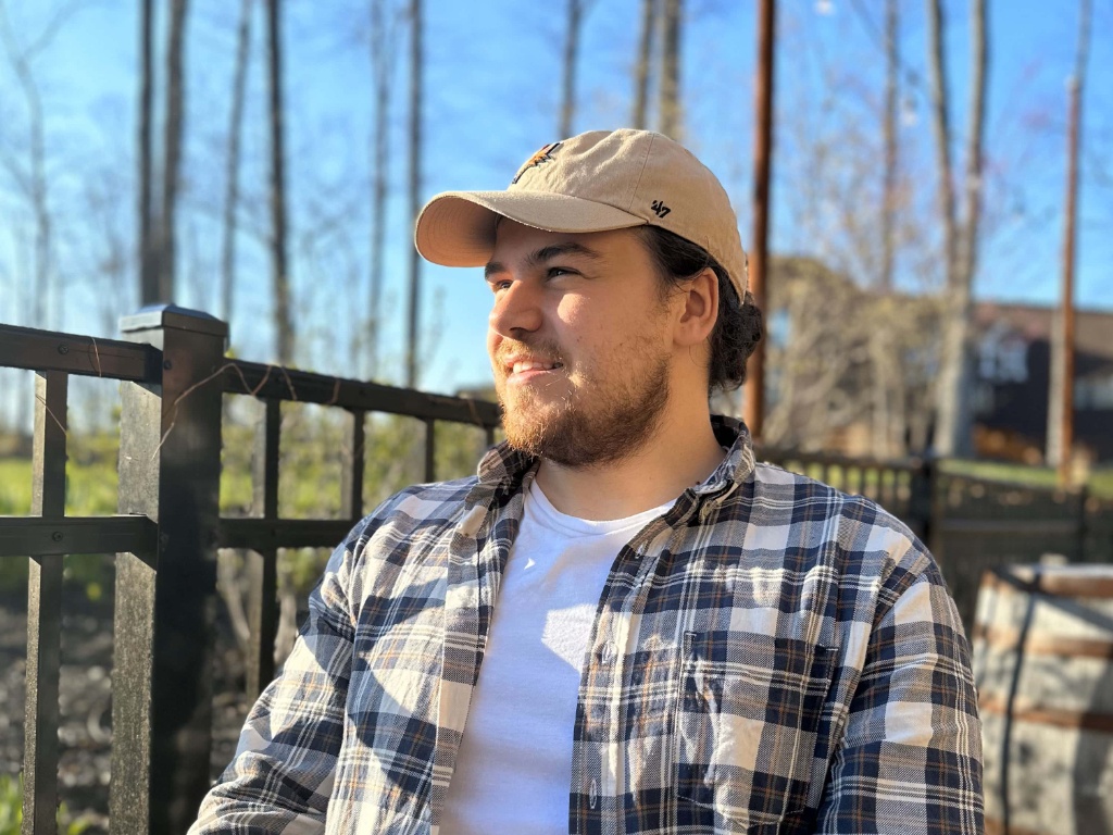 A young man in a plaid shirt with a baseball cap sits on a deck and looks off to the left