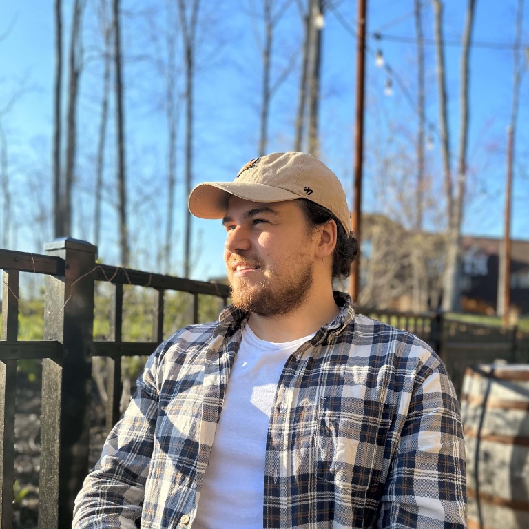 A young man in a plaid shirt with a baseball cap sits on a deck and looks off to the left