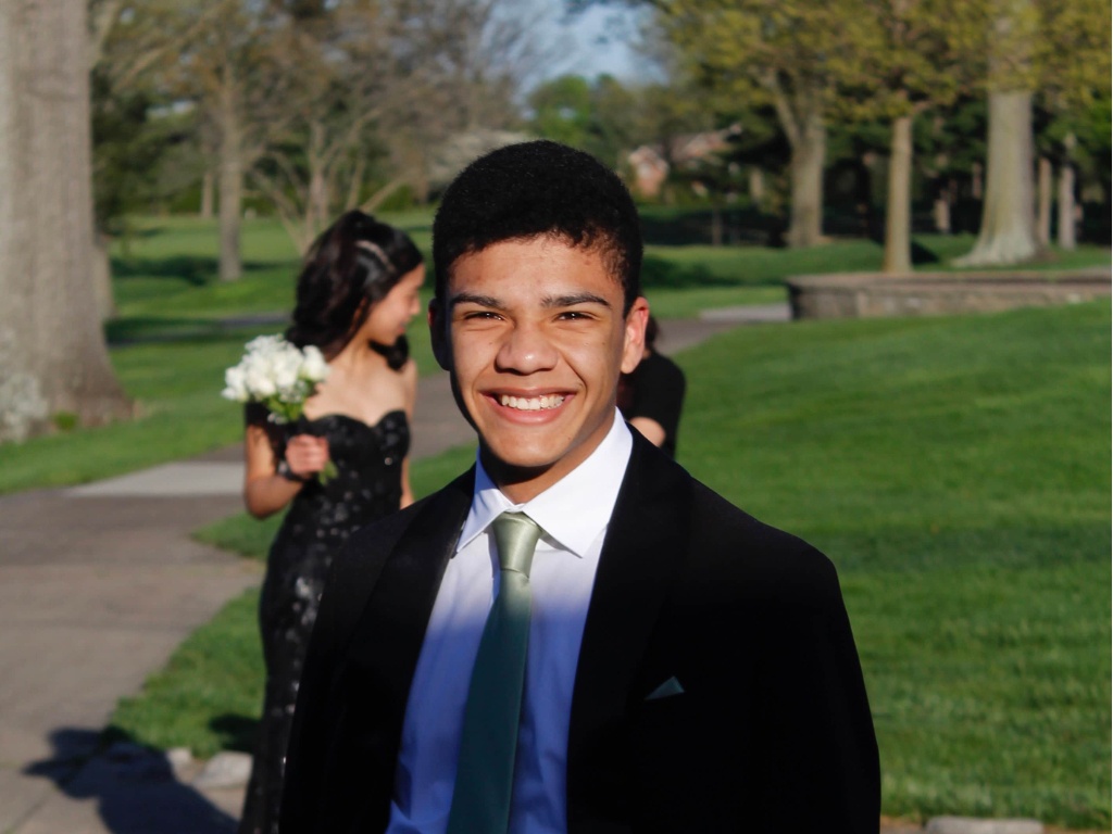 A young man in a suit poses for prom pictures in a green area on a sunny day