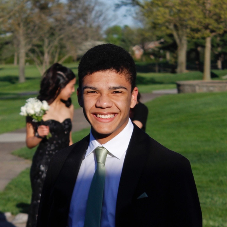 A young man in a suit poses for prom pictures in a green area on a sunny day