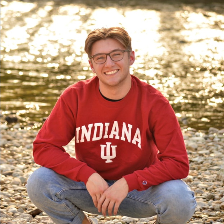 A young man in an IU sweatshirt squats in front of a body of water while smiling at the camera