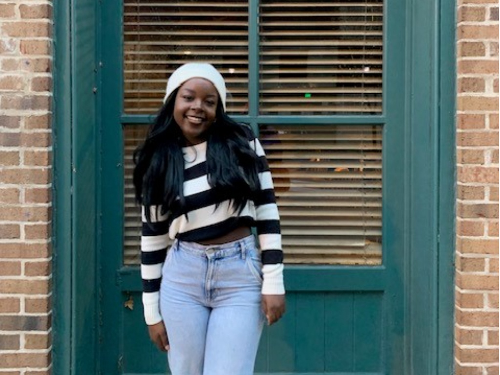 A young woman poses in a striped shirt and white hat in front of a green door