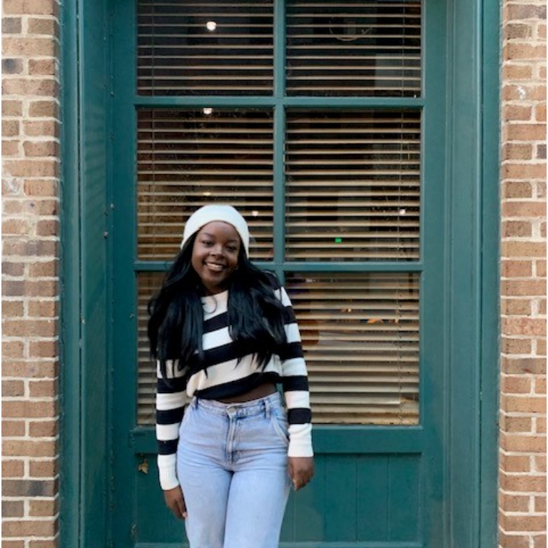 A young woman poses in a striped shirt and white hat in front of a green door