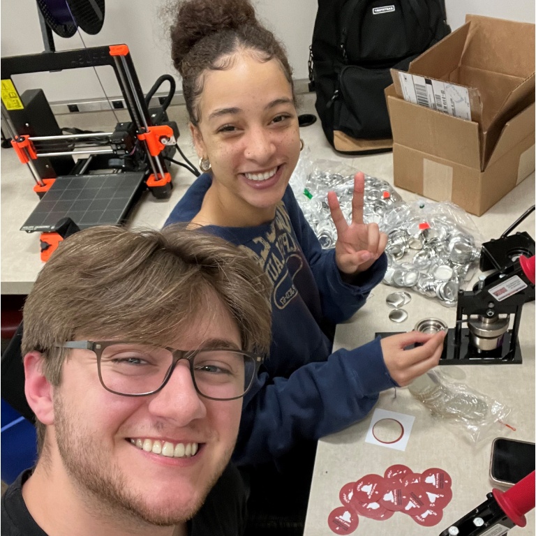 Two students pose for the camera while making pin-on buttons at Wells Library