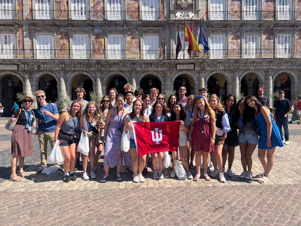 A group of students stands together in front of an older looking building while holding an IU flag