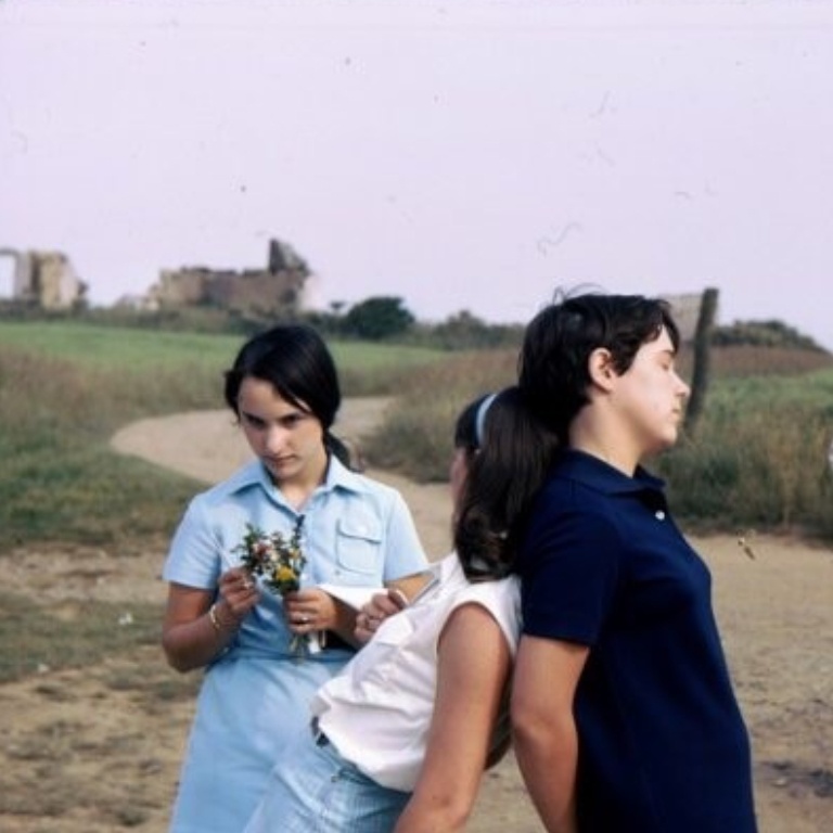 Two students lean against one another back-to-back while another to the left holds a small bouquet of wildflowers 